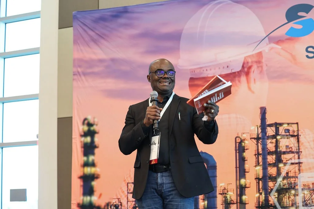 A man with glasses in a black blazer and jeans speaking into a microphone while holding a booklet, standing in front of a colorful backdrop with industrial structures and a sunset skyline.