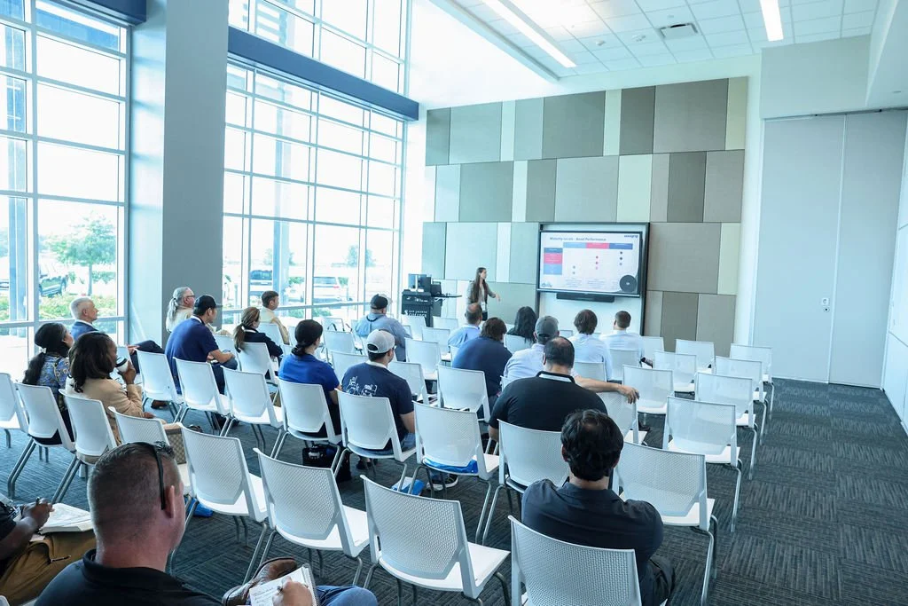 A diverse group of people attending a presentation in a modern conference room with large windows, a presenter standing near a large screen displaying a chart or graph.
