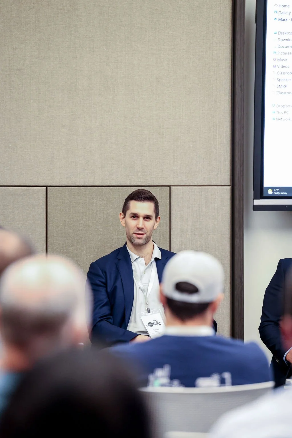 A man in a dark blazer and white shirt sitting in front of a beige wall during a discussion or presentation at a conference, with an audience in the foreground.