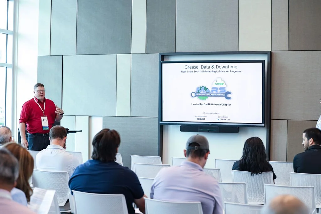 A man in a red shirt giving a presentation at a conference with an audience seated facing a large screen. The screen displays the title "Grease, Data & Downtime" and logos related to maintenance and lubrication.
