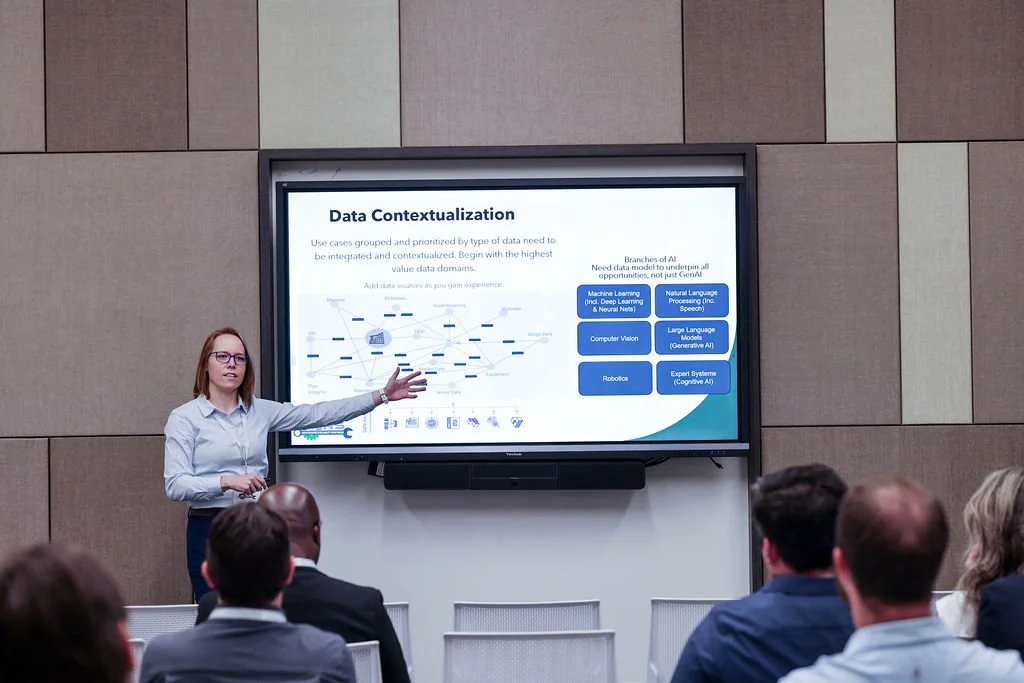 A woman giving a presentation on data contextualization to an audience in a conference room. She is pointing at a large screen displaying a diagram with blue text and boxes.