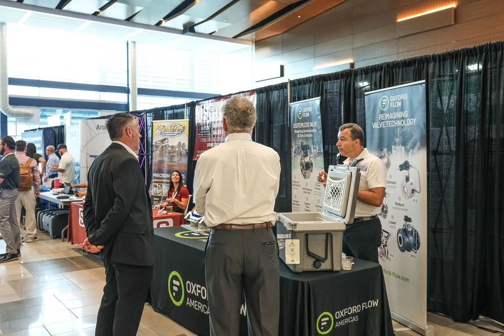 Business professionals at a trade show discussing at an Oxford Flow booth with banners displaying valve technology.