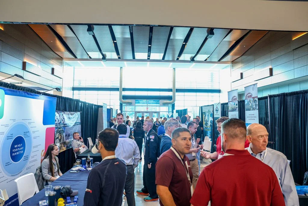 A busy conference with attendees interacting and networking in a large, well-lit hall with display booths and informational banners.