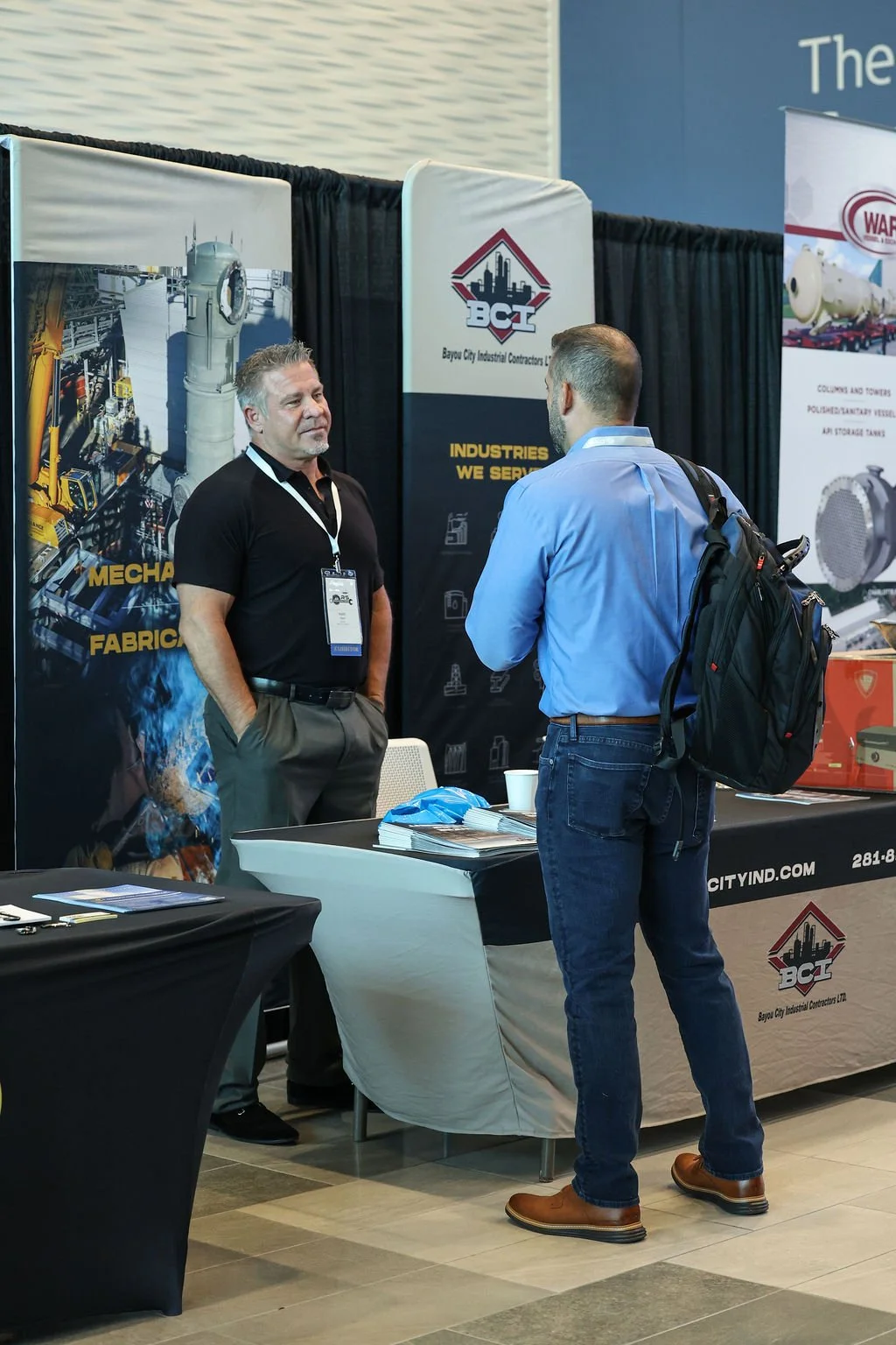 Two men talking at an industrial trade show booth. One man, with gray hair, wearing a black shirt and gray pants, is standing behind the booth. The other man, with a beard, wearing a blue shirt and khakis, is facing him with a backpack. The booth has