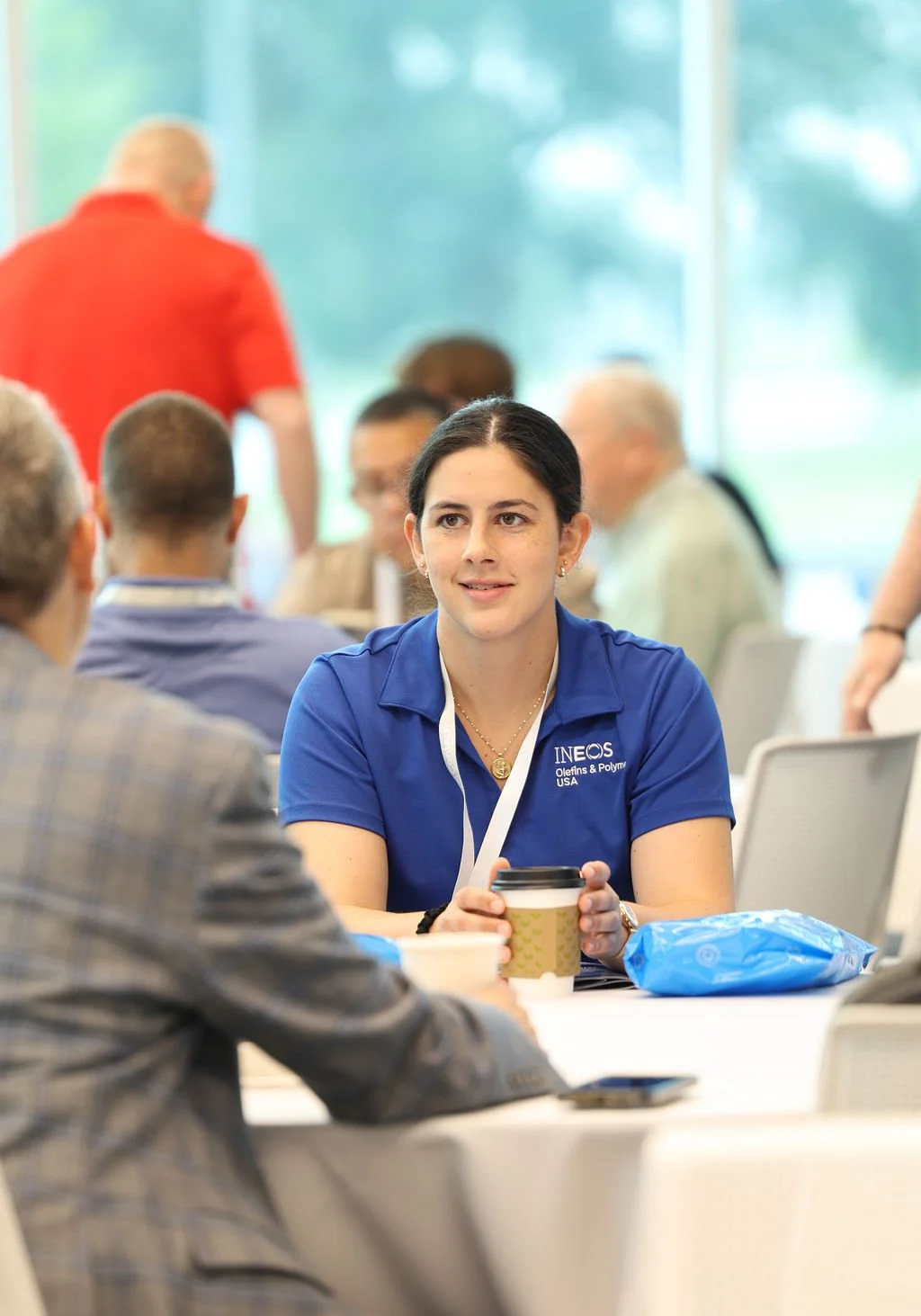 A young woman with dark hair, wearing a blue polo shirt with the INEOS logo, sitting at a table during a conference or event, holding a disposable coffee cup and listening attentively.