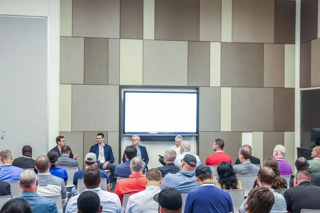 A conference or panel discussion taking place in a large room with a diverse audience, four men are seated at the front panel, with one standing and speaking, and a large screen displaying a presentation behind them.