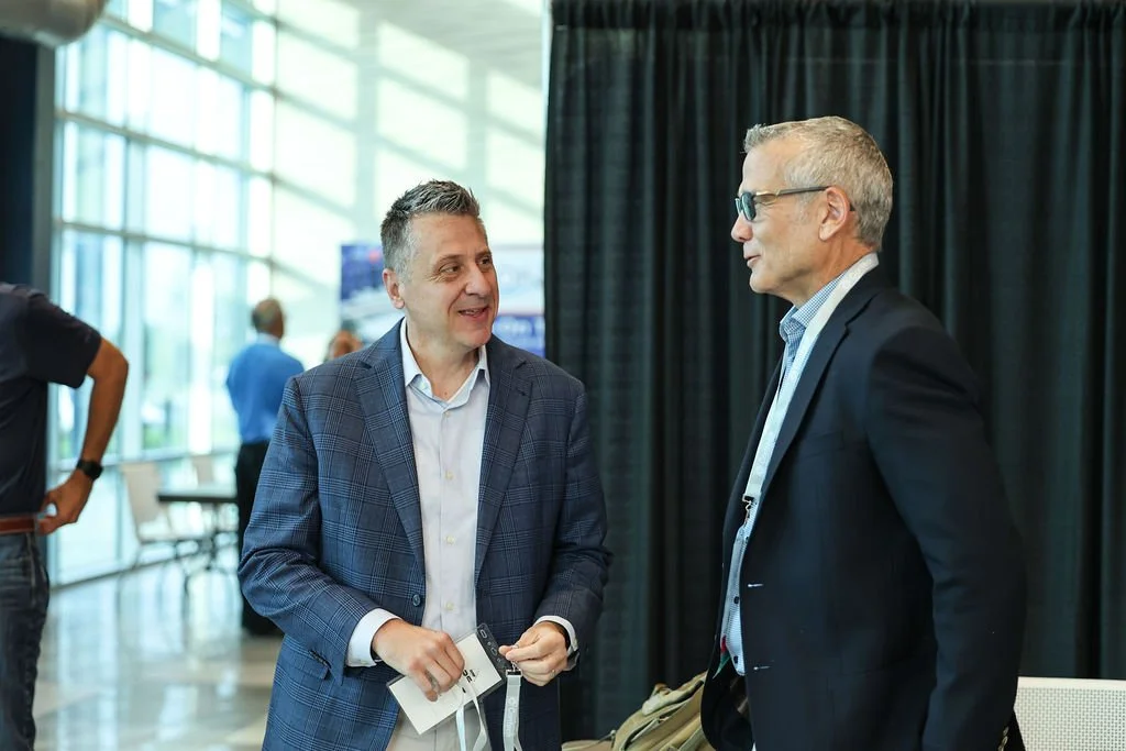 Two men in business suits talking at a professional event, with large windows and a black curtain in the background.