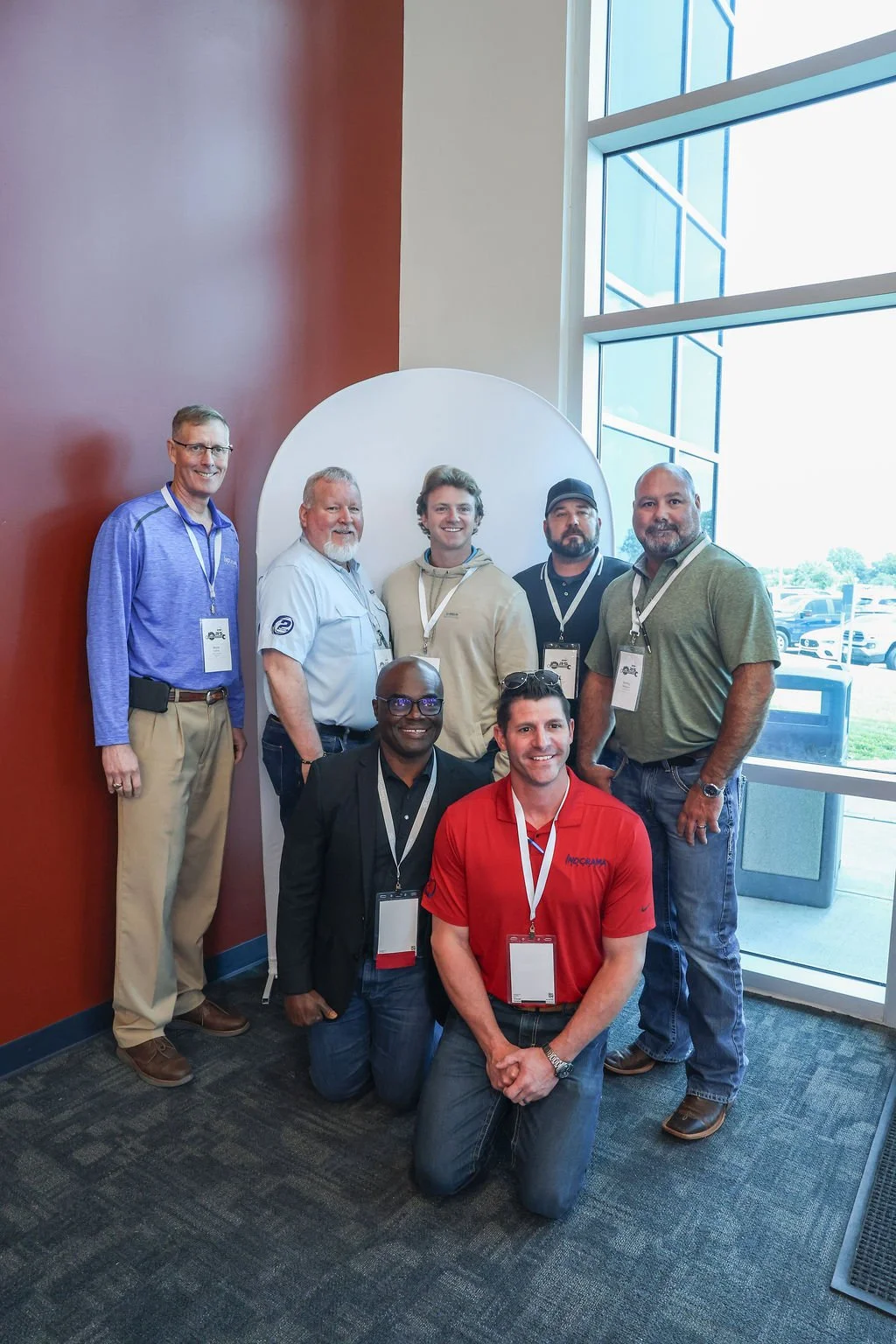 A group of seven men posing for a photo in a conference setting, some wearing conference badges, standing near a large window with a view of a parking lot.