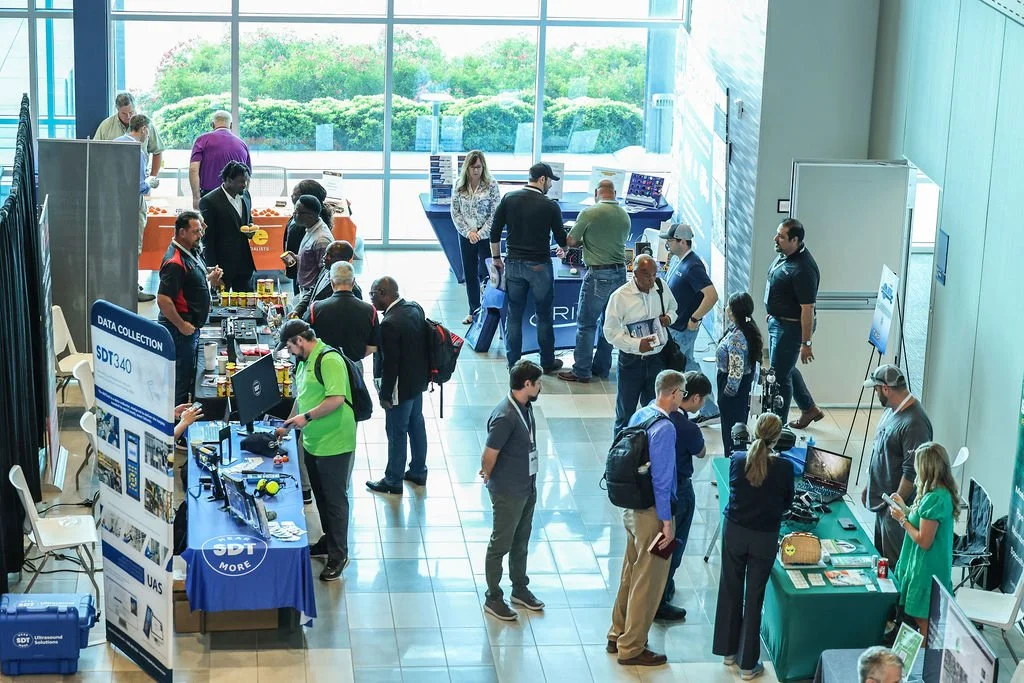 People attending a technology trade show or conference in a large, well-lit atrium with floor-to-ceiling windows showing greenery outside. Several booths display electronic devices, and attendees are engaging with exhibitors and exploring products.