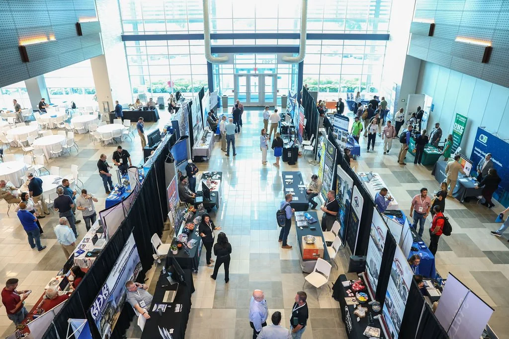 A busy convention center with multiple exhibitor booths displaying technology and promotional materials, and attendees walking around and engaging with vendors.