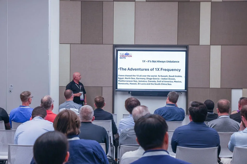 A man is giving a presentation to an audience in a conference room. The screen displays the title "The Adventures of 1X Frequency" and a list of locations the speaker has traveled to.