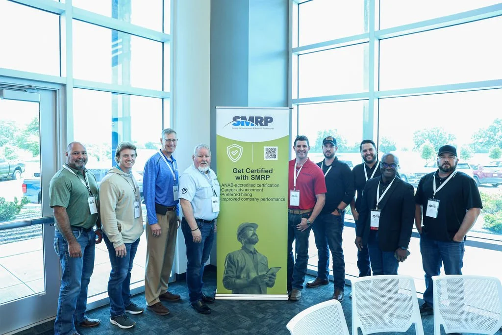 Group of nine people standing in a conference room with floor-to-ceiling windows, holding a banner for SMRP, a certification organization.