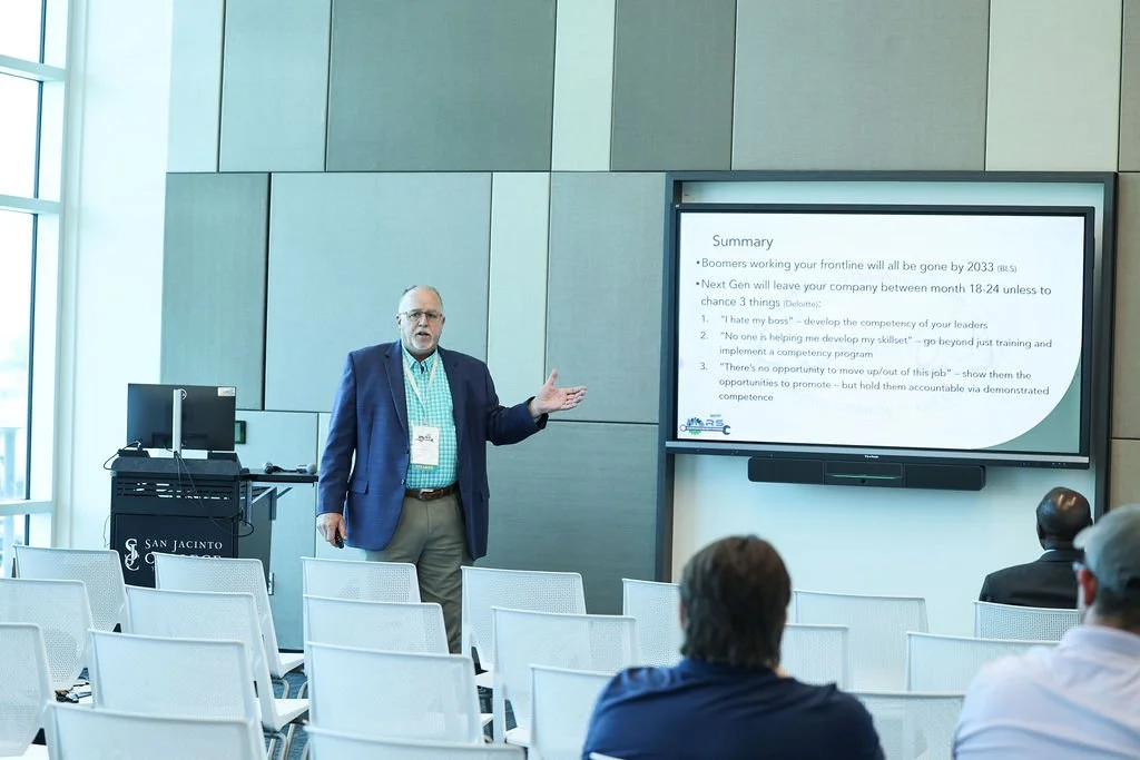A man in a blazer and khakis standing in front of a large digital screen presenting to an audience in a modern conference room.