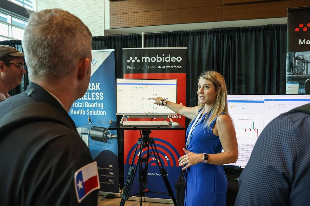 A woman in a blue dress shows data on a screen at a technology exhibition, with banners behind her displaying 'mobideo' and 'digitalizing the industrial workforce'. Several attendees are watching her presentation.