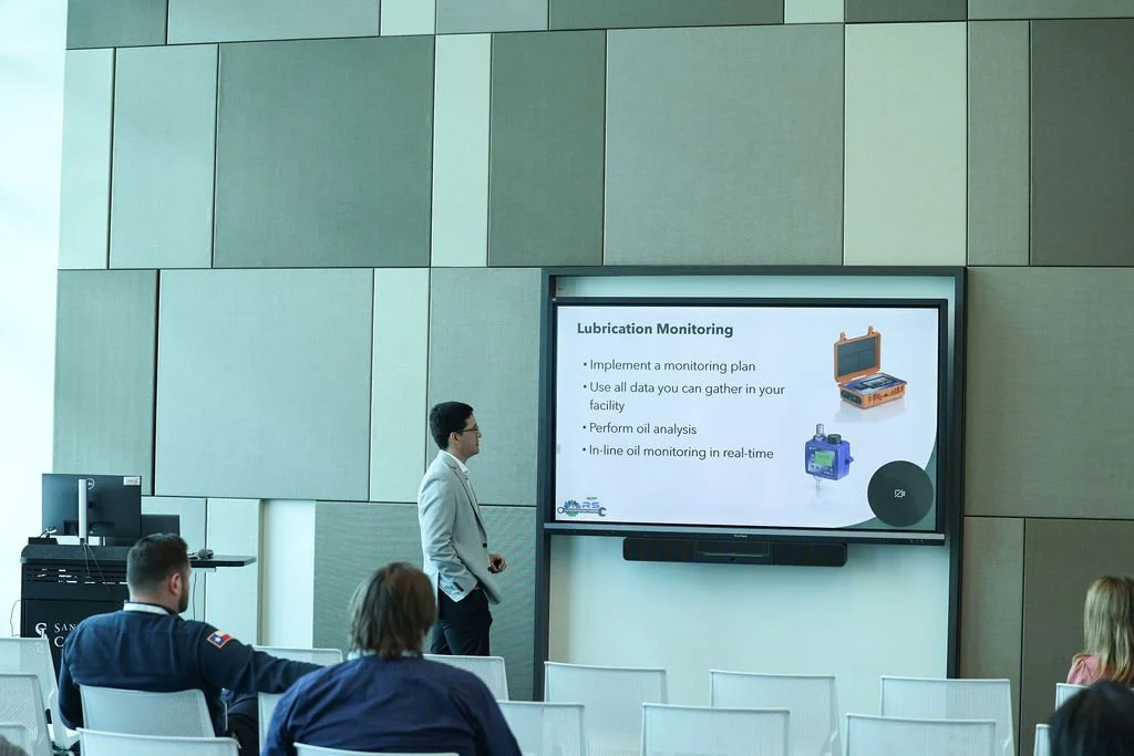 A man giving a presentation on lubrication monitoring to an audience in a conference room, with a slide showing bullet points and images of monitoring equipment.