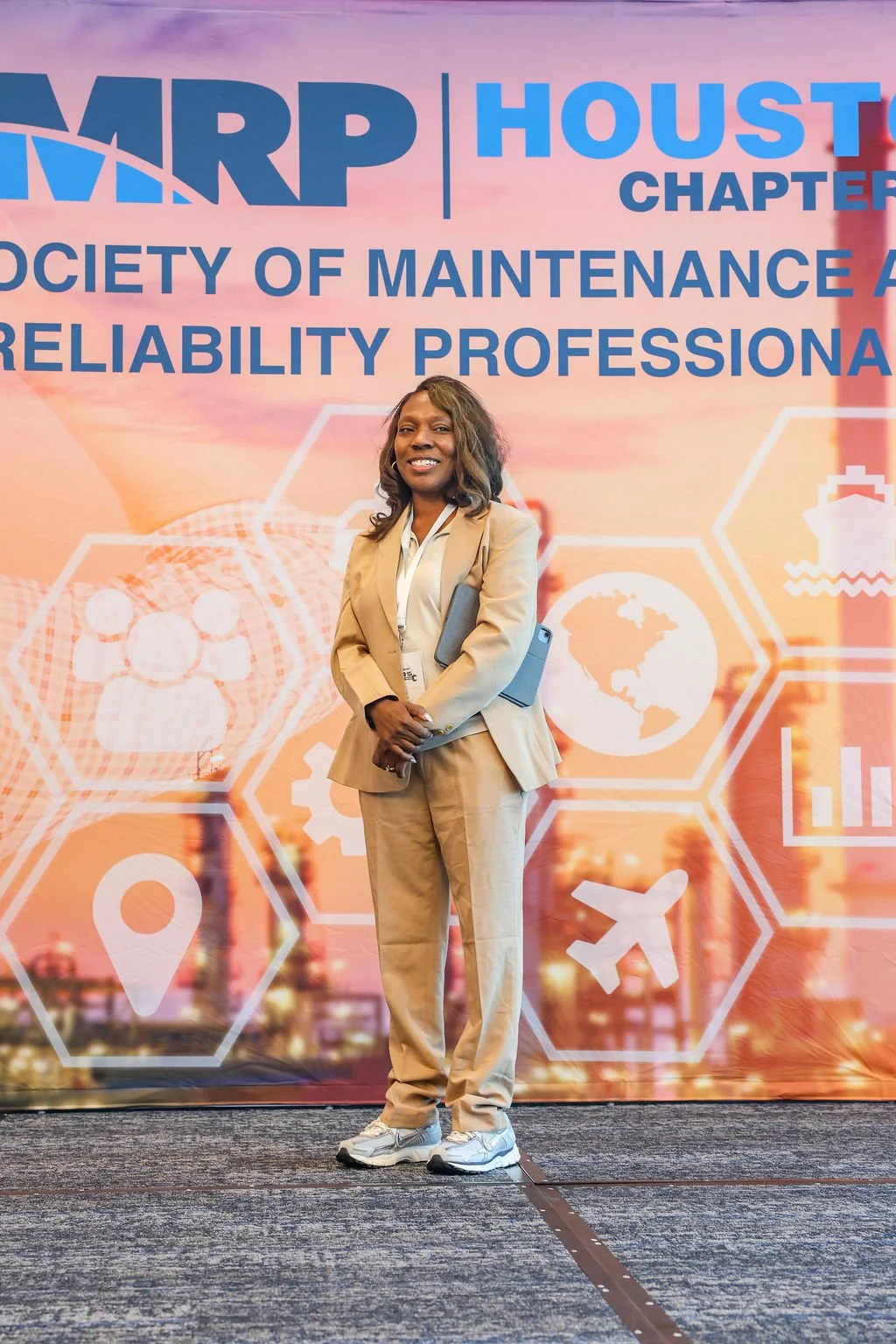 A woman in a beige suit holding a folder stands in front of a conference backdrop with the words "HARP Houston Chapter Society of Maintenance & Reliability Professionals" and various icons related to transportation, logistics, and global connectivity