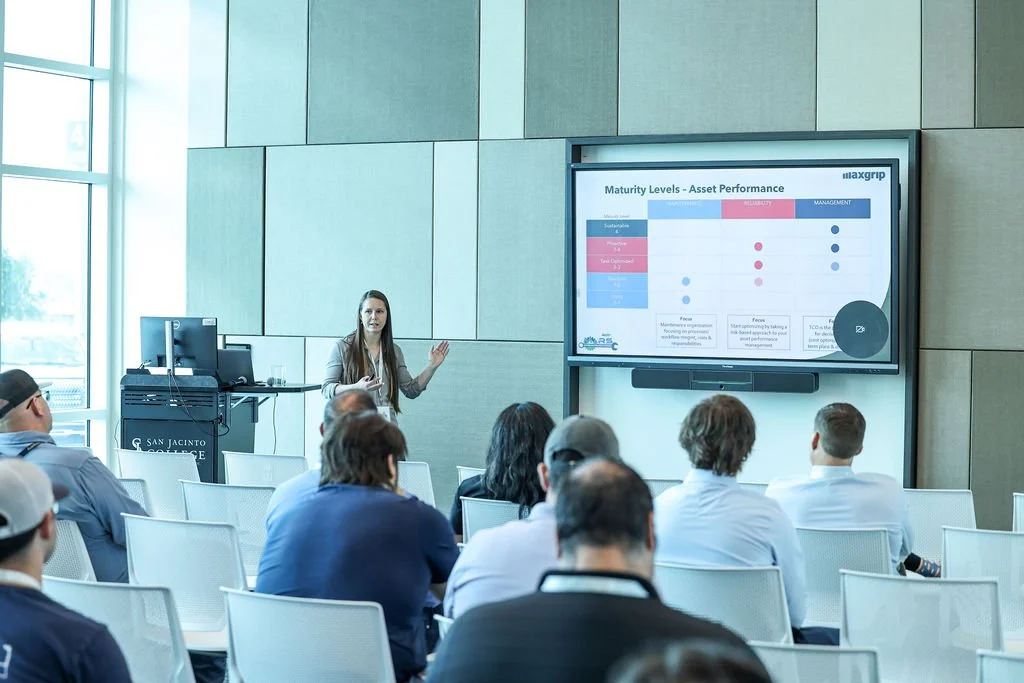 A woman giving a presentation to a group of people in a conference room at San Jacinto College. The presentation slide shows a chart with maturity levels and asset performance.