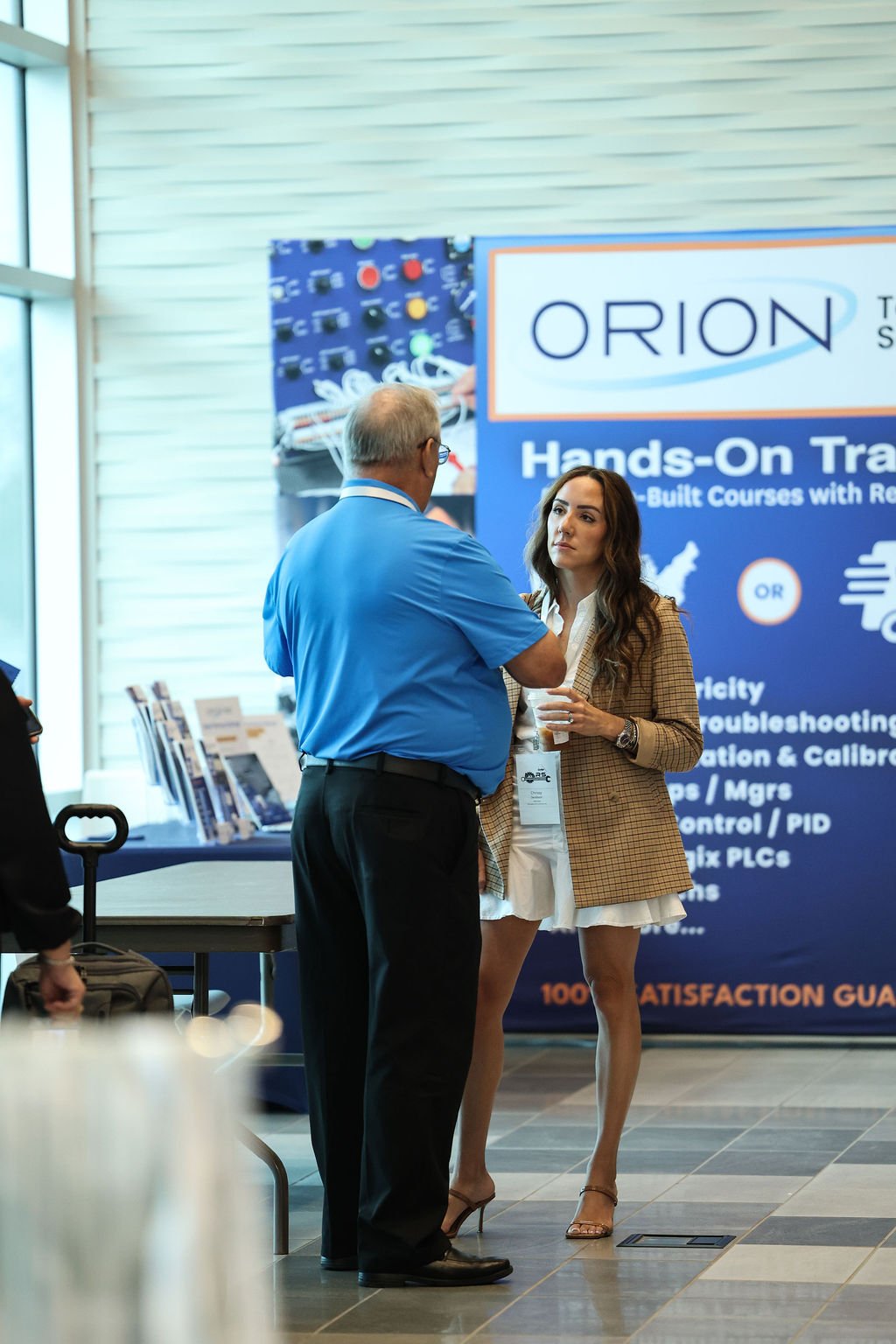 A man and a woman having a conversation at a conference booth for Orion Training, with a blue poster display behind them.