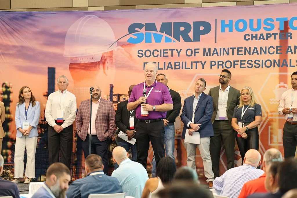 A group of professionals standing on stage during a conference, with a large banner behind them displaying 'SMRP Houston Chapter' and 'Society of Maintenance and Reliability Professionals'. Some are holding papers or microphones, and audience members
