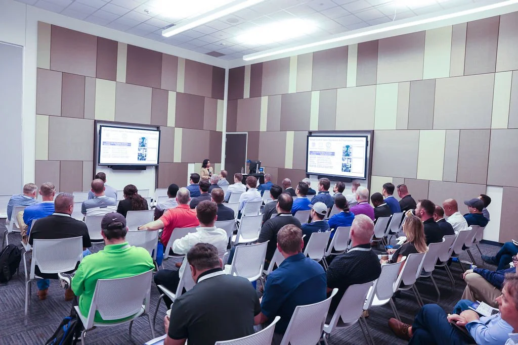 Conference room filled with attendees watching a presentation on two wall-mounted screens.