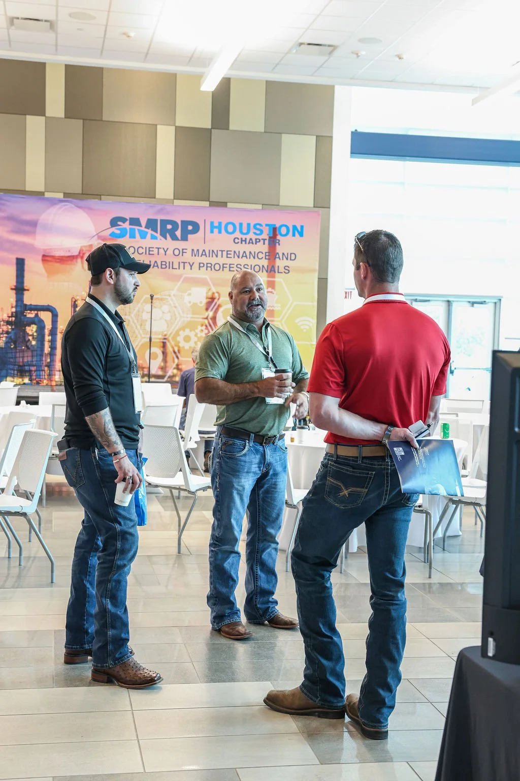 Three men engaged in conversation at an indoor conference with a banner for the Society of Maintenance and Reliability Professionals (SMRP) Houston Chapter in the background.