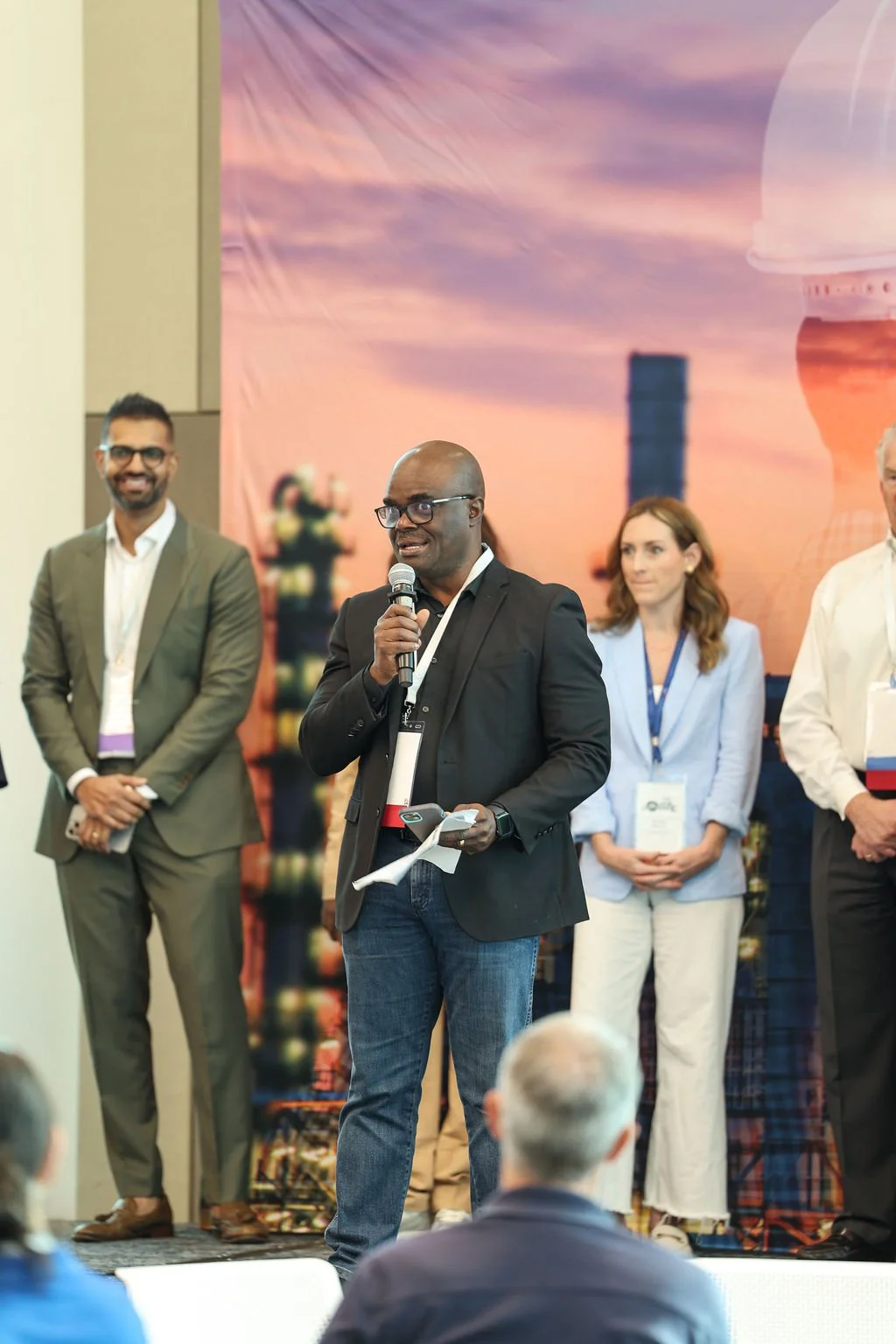A man in a black blazer speaking into a microphone at a conference, with several people standing behind him against a backdrop featuring a cityscape and a sunset sky.