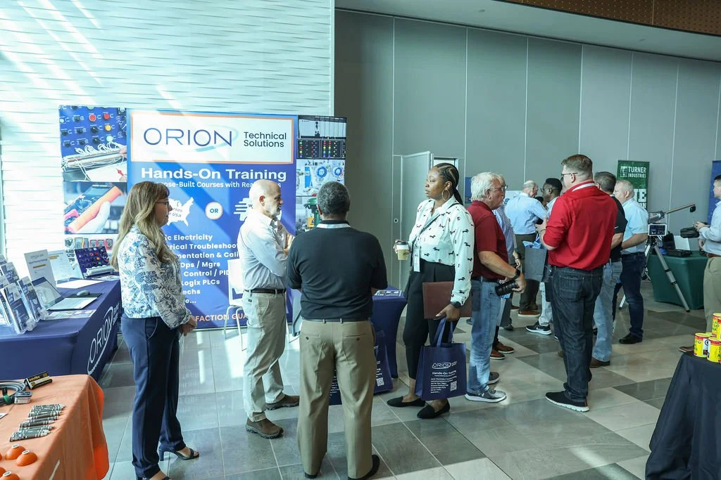 People attending a technical training event at Orion Technical Solutions booth, engaged in conversations, with various informational displays on the table.