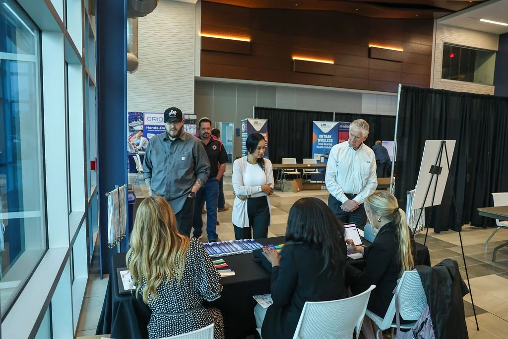 People at an exhibition booth inside a conference center, engaging in discussions near display tables, with promotional banners and a black curtain backdrop.