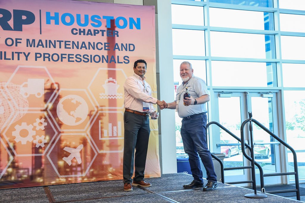 Two men shaking hands on stage at Houston chapter event, one holding a bottle, with a large banner behind them displaying the words 'Houston Chapter' and icons representing logistics and maintenance.
