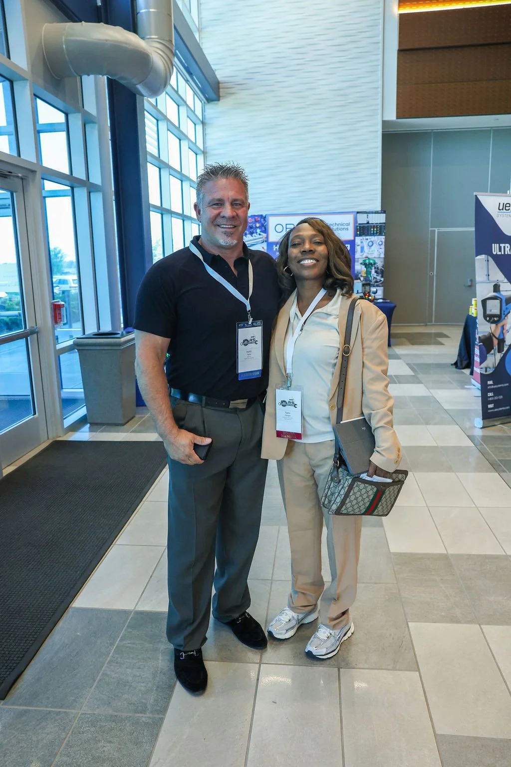 A man and woman smiling inside a conference center, wearing badges, standing in front of display booths.