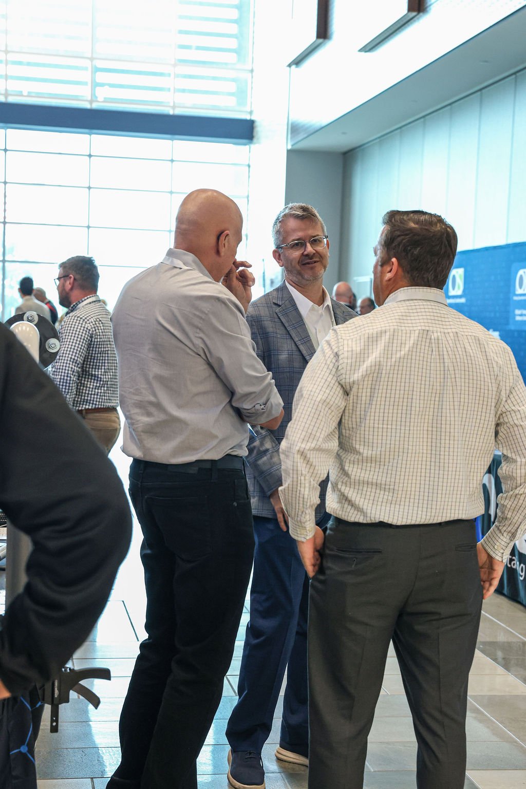 Three men engaged in conversation at a professional event or conference, with a large window and others in the background.