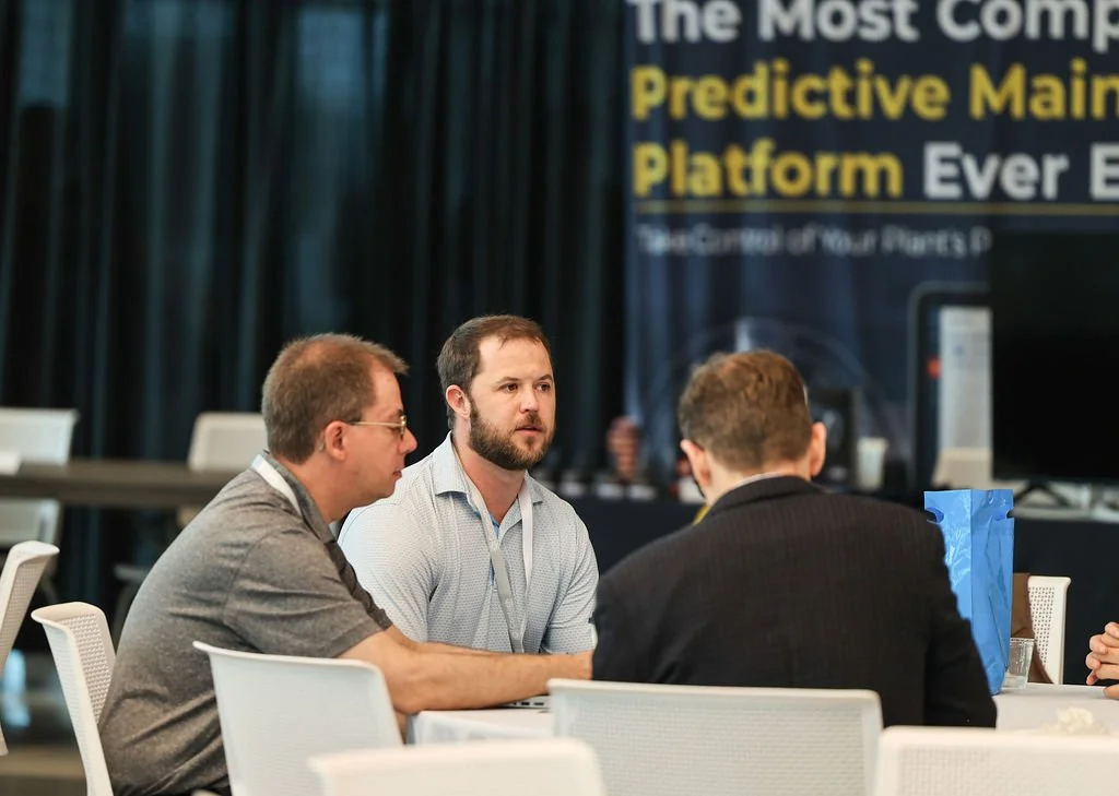 Three men sitting at a conference table, engaged in conversation, with a large display screen in the background showing text about predictive mainframe platform.