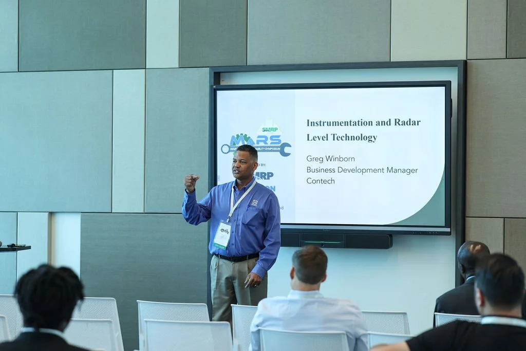 A man giving a presentation at a conference, standing in front of a large screen that displays the title "Instrumentation and Radar Level Technology" and his name, Greg Winborn, Business Development Manager at Contech. The audience is seated, listeni