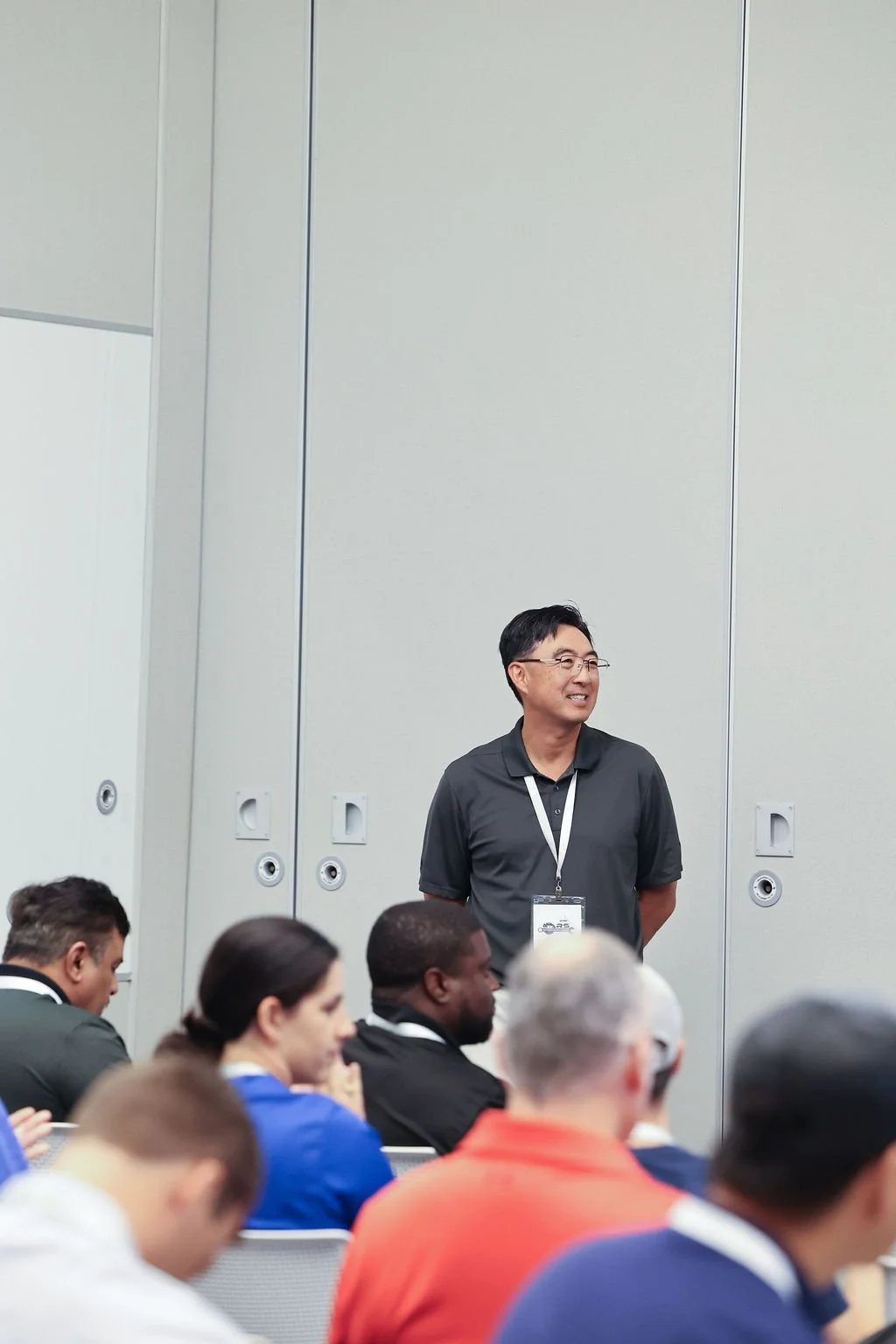 Man standing and smiling at a conference, wearing glasses, a black polo shirt, and a conference badge, with an audience seated in front of him.