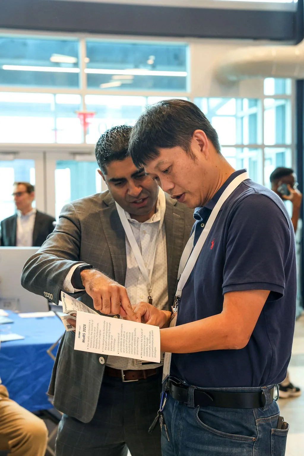 Two men are looking at a brochure or flyer together at what appears to be a conference or event. One man is wearing a suit, and the other is in a polo shirt. They are indoors with large windows in the background.