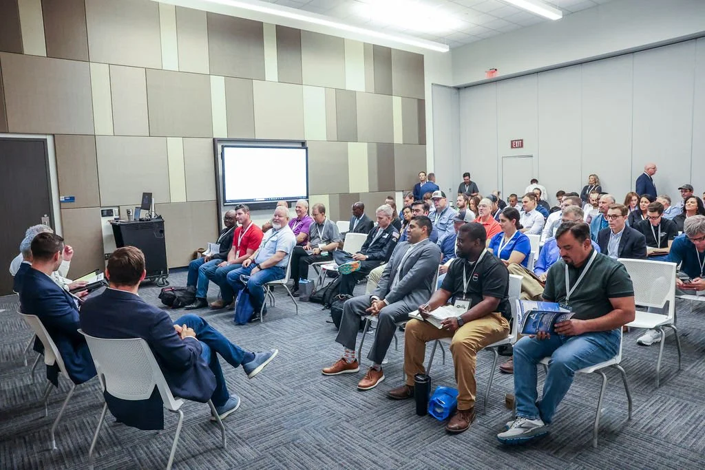 A diverse group of people attending a conference or seminar in a modern conference room, seated in rows facing a panel or speaker area.