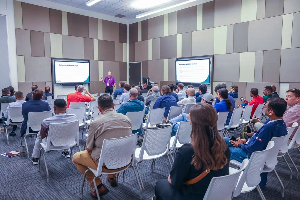 A conference room filled with diverse attendees watching a speaker presenting at the front. Two large screens display presentation slides.