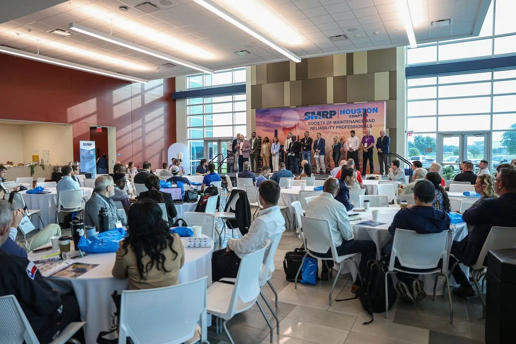 Attendees seated at round tables listening to a group of speakers on stage during a conference at the SMART Houston Chapter of the Society of Maintenance and Reliability Professionals.