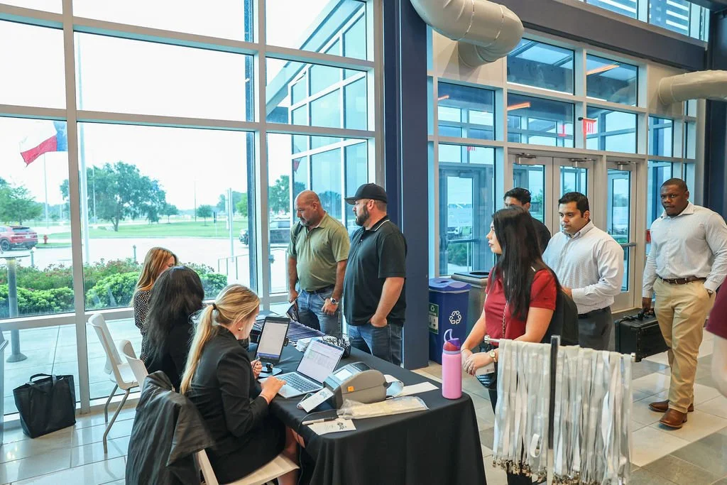 People standing in line at a registration desk inside a modern building with large glass windows showing a parking lot and trees outside.