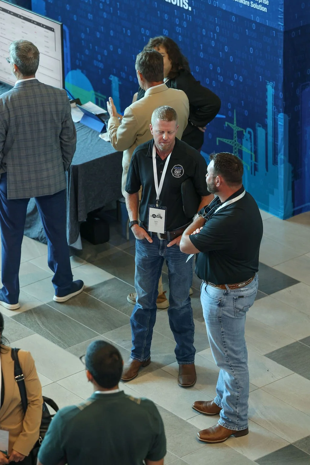 Group of people at a conference or trade show engaging in conversation, with some standing near a booth with digital and technological graphics on the background.