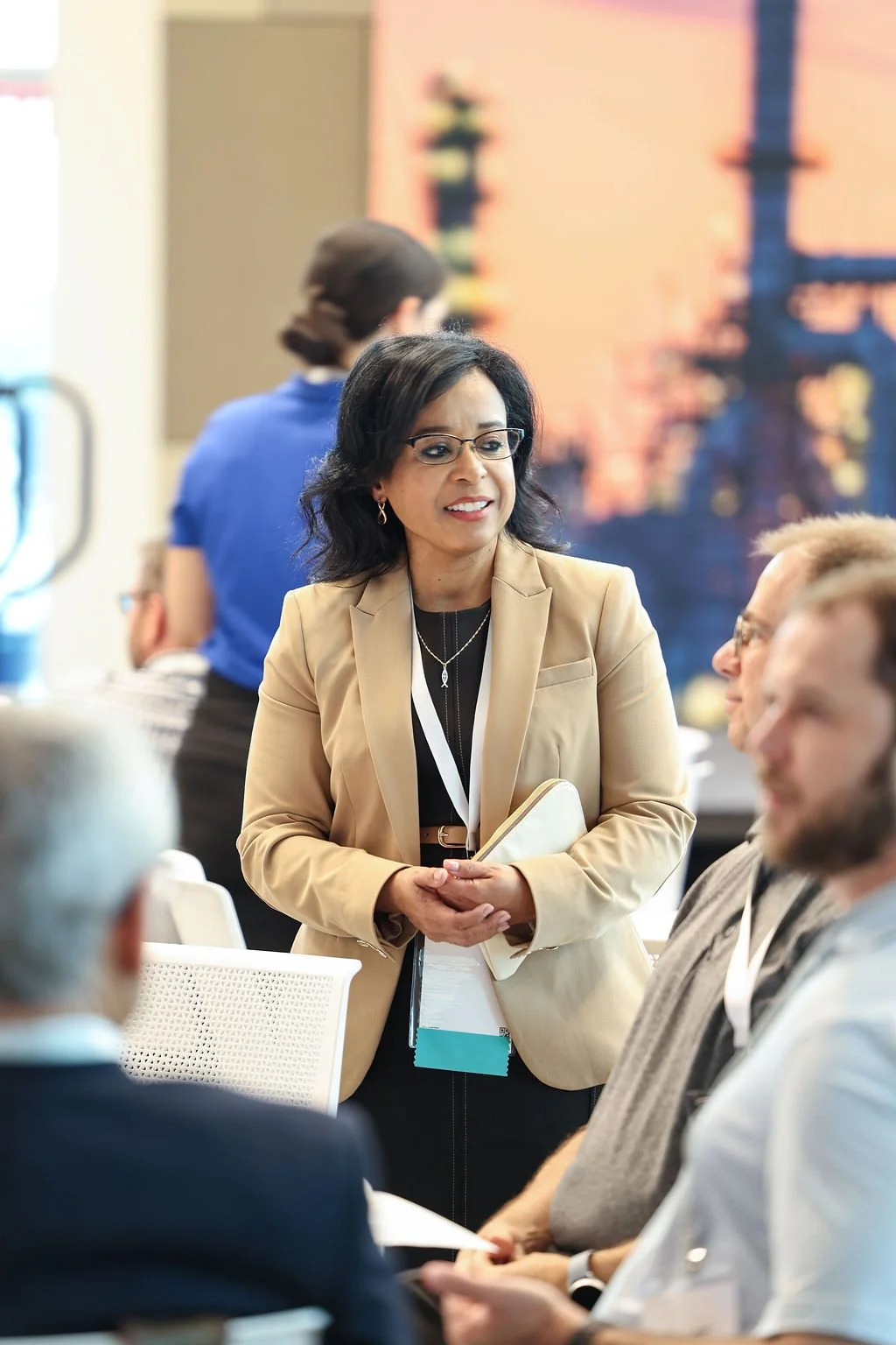 A professional woman in a beige blazer and glasses engaging in conversation at a business meeting, with other seated participants and a large cityscape image in the background.