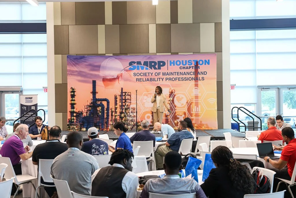 A woman on stage speaking at the Society of Maintenance and Reliability Professionals (SMRP) Houston Chapter conference, with an industrial background banner and an audience seated at round tables.