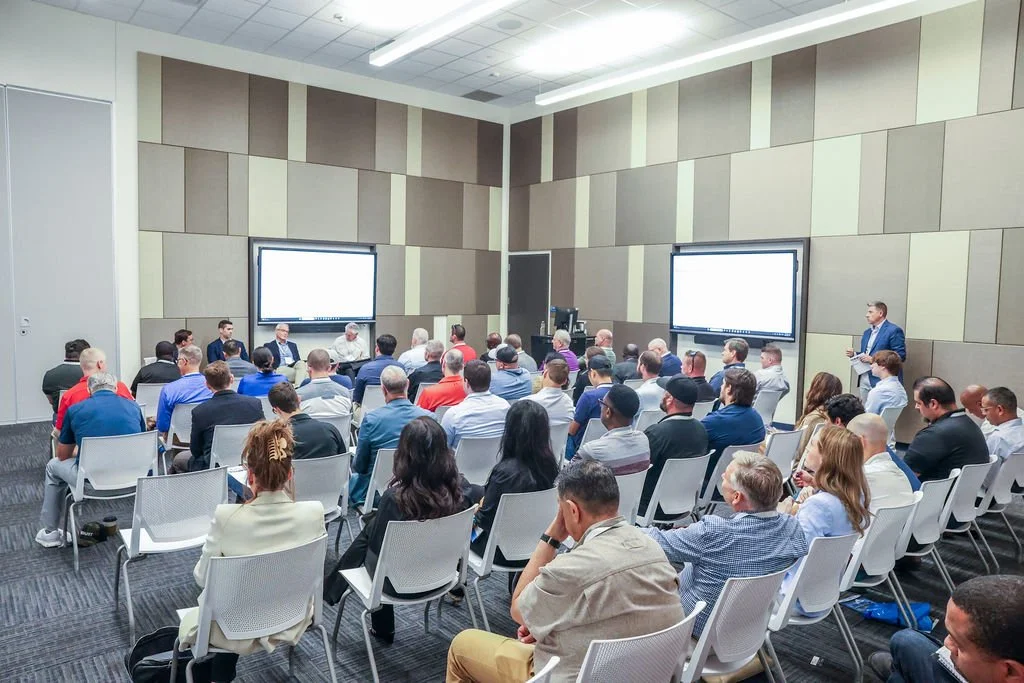 A large conference room filled with people attending a presentation. There are two large screens at the front, and a panel of speakers seated at a long table.