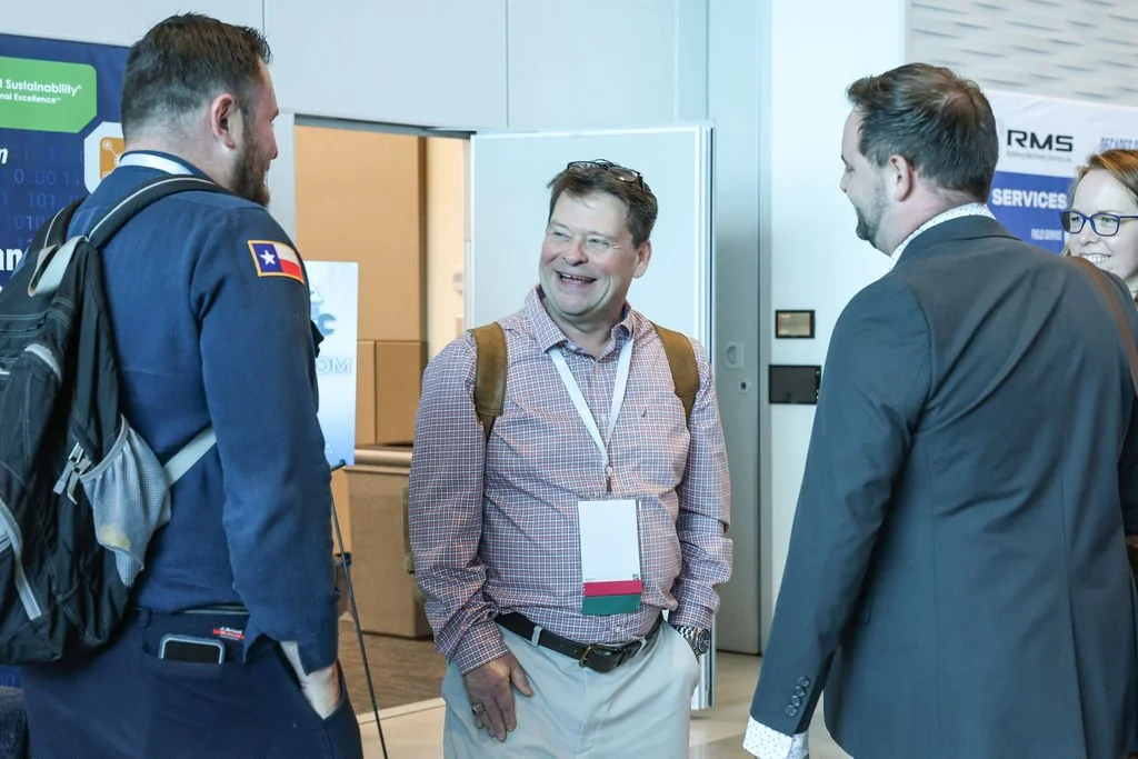Four people, three men and one woman, are engaged in conversation at a conference. They are smiling and wearing name badges. The man on the left has a backpack, the man in the middle is wearing a checkered shirt, and the man on the right is in a suit