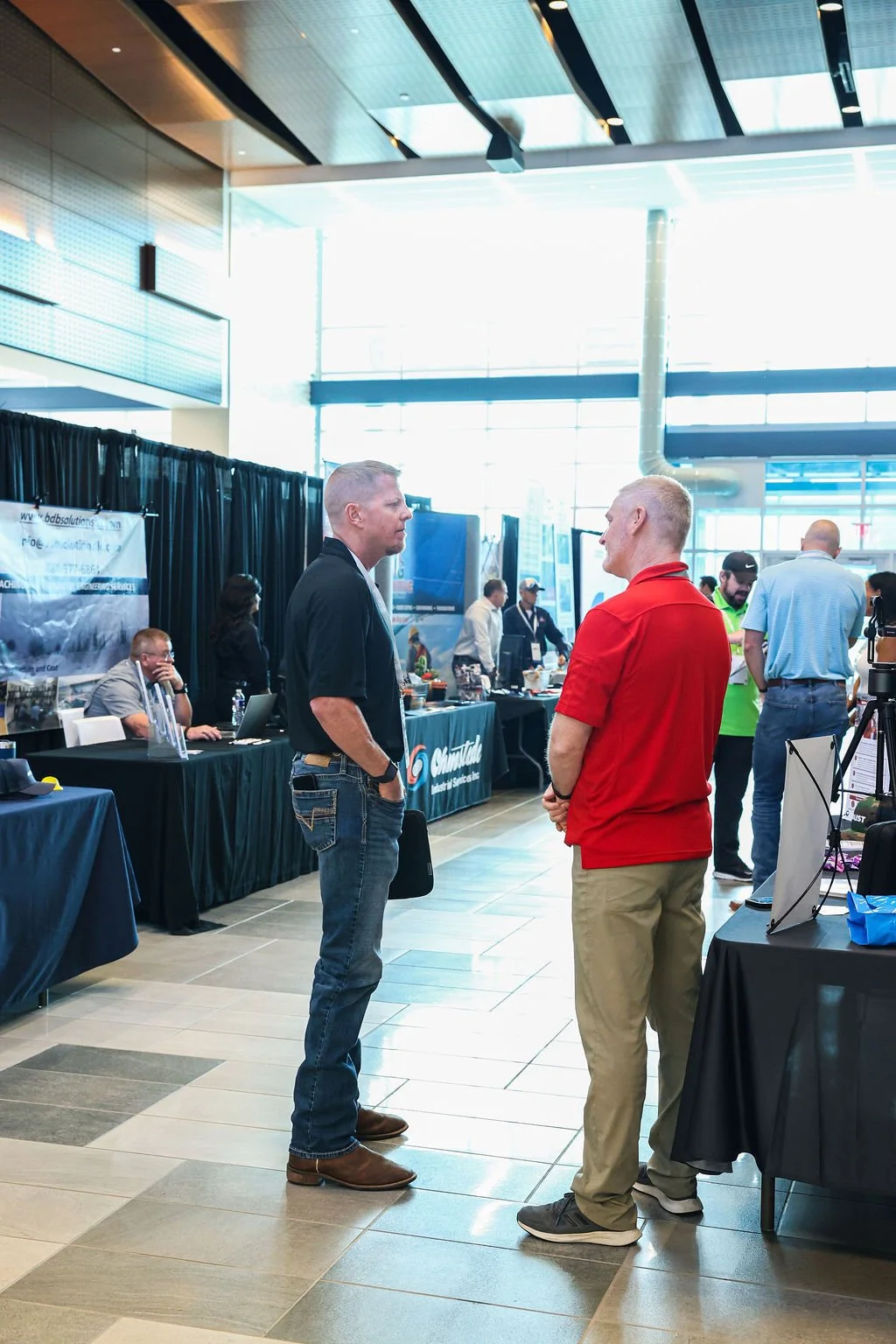 Two men are talking at a business conference or trade show. They are standing indoors near exhibition booths with displays and people working behind tables. One man is wearing a black shirt and jeans, and the other is wearing a red polo shirt and kha