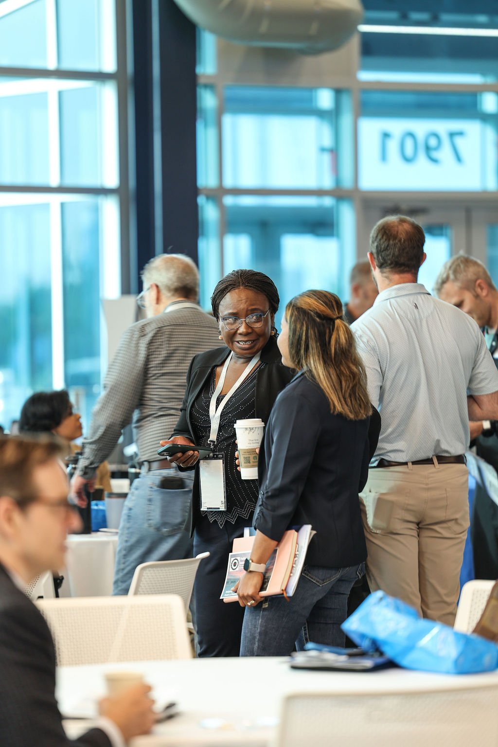 People engaging in conversation at a professional conference or event in a modern, glass-enclosed indoor space.