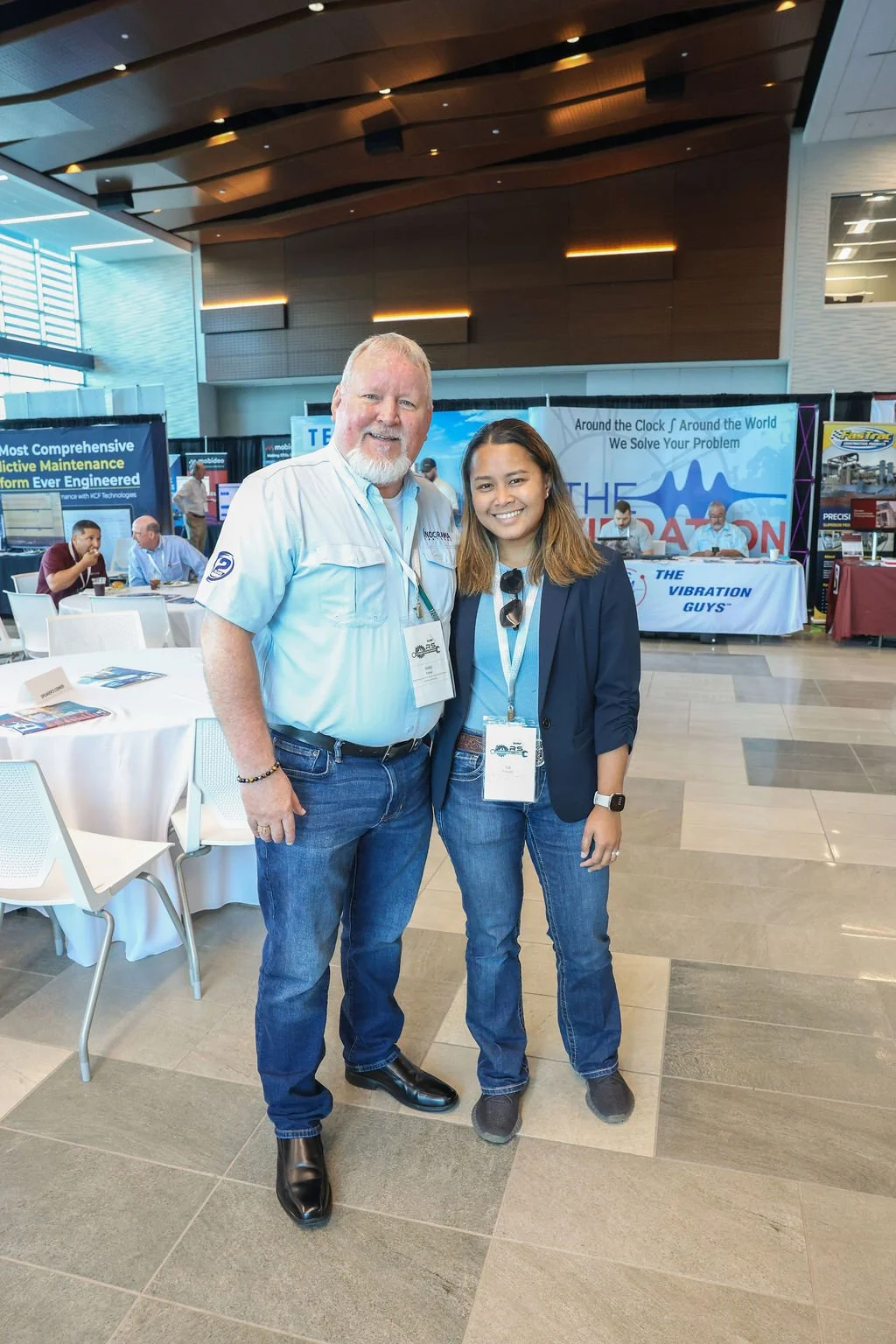 Two smiling people standing together inside a conference or expo hall, with booths and banners in the background. The man on the left has white hair and beard, wearing a light blue shirt and jeans. The woman on the right has brown hair, wearing a bla