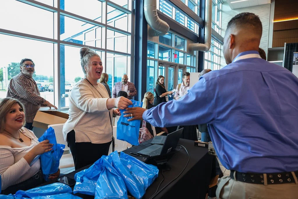 A woman in a white jacket is smiling as she receives a blue gift bag from a man in a blue shirt at a conference or event. There are other people in the background, some holding similar bags, inside a room with large windows and modern architecture.