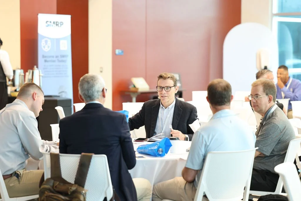 Six men in business casual attire sitting around a table engaged in a discussion at a conference or meeting. Conference room with a banner in the background.
