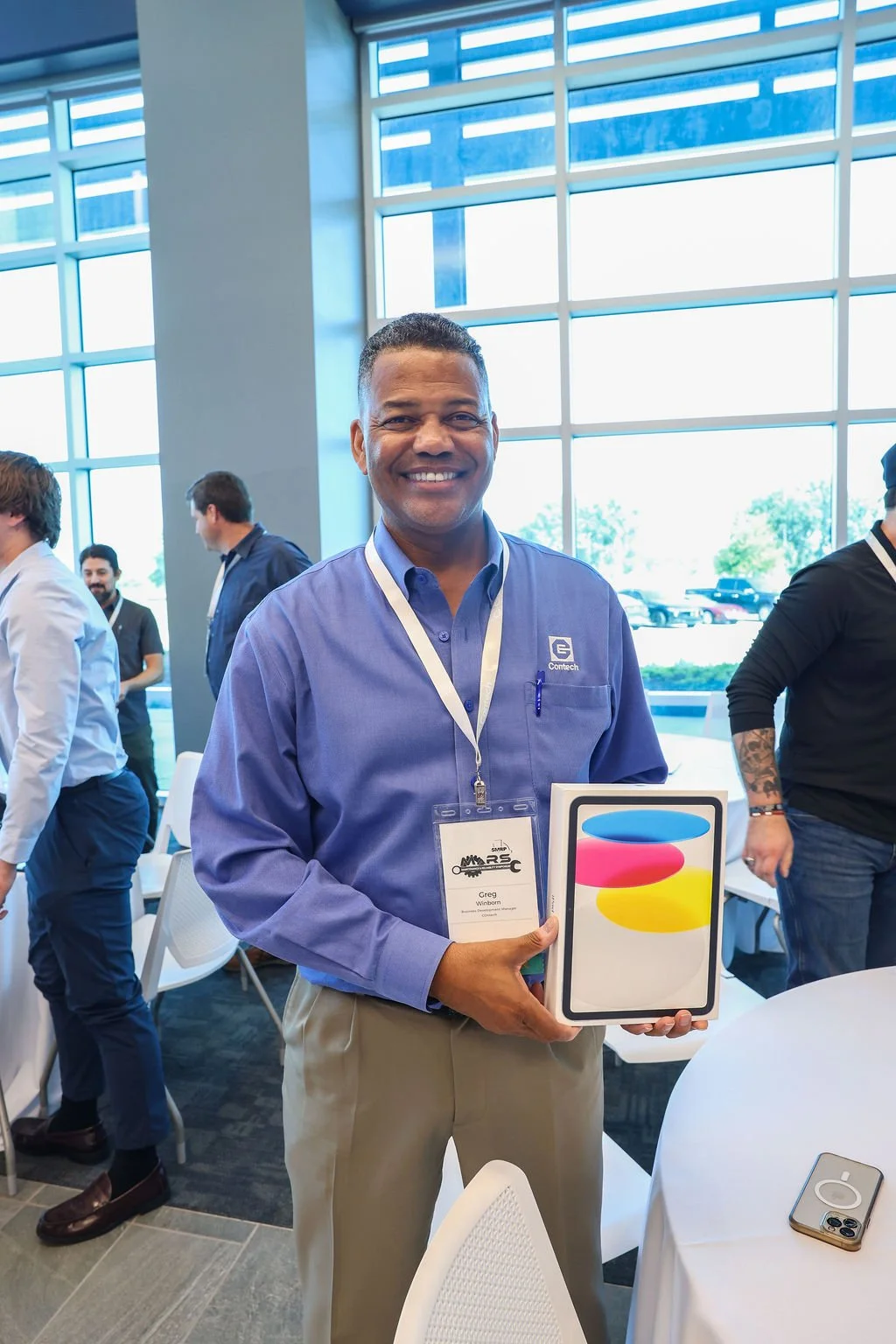 A smiling man in a blue shirt holding a colorful box at an indoor event with large windows and other people in the background.